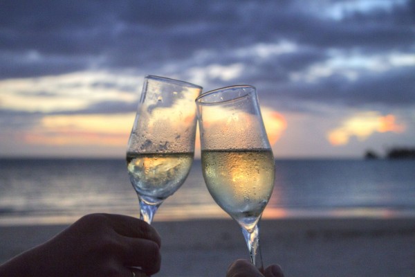 Two wine glasses clink together in front of a beach sunset